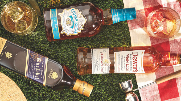 Bottles of blended scotch—Johnnie Walker Blue Label, Dewar's White Label, and Chivas Regal Mizunara—rest on their sides, pictured from a bird's-eye view, atop what appears to be close-cropped grass, next to a red and white plaid-patterned picnic blanket, along with a wooden-handled metal jigger, and two rocks glasses containing ice and what appears to be whisky cocktails; the brim of what appears to be a straw hat also is partially in view to the lower left corner of the photo.