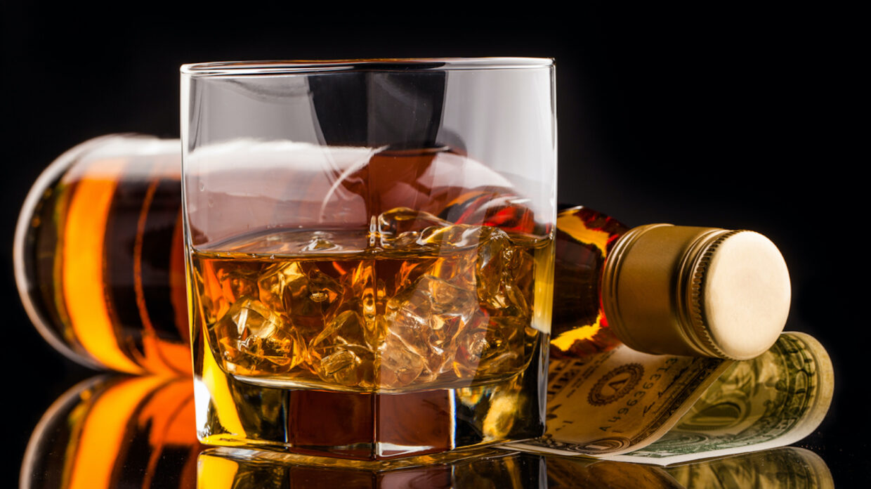 glass of whisky with bottle on reflective surface with black background and dollar money