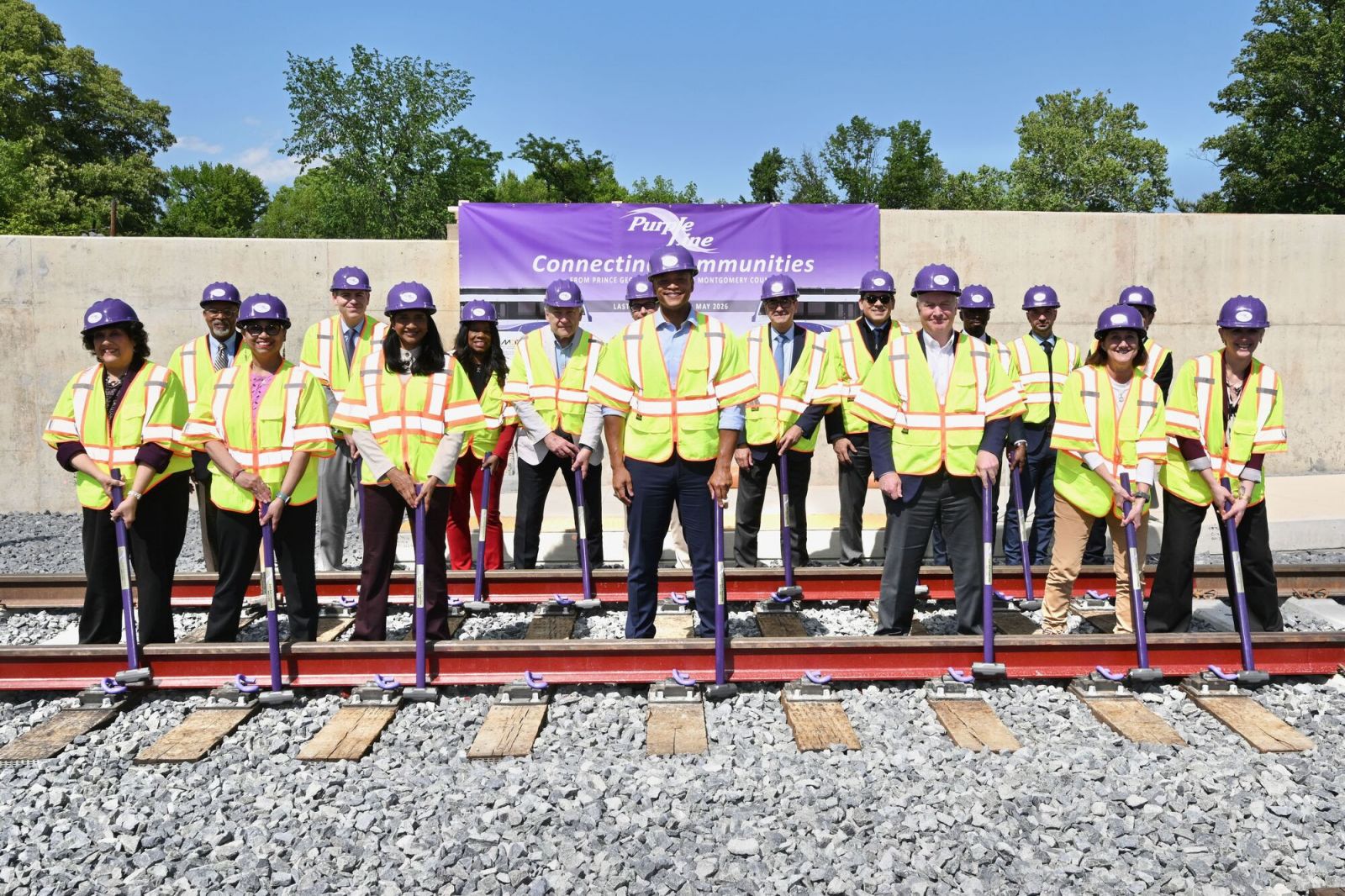 The Governor and other officials, dressed in high visibility vests and holding sledgehammers, stand along railroad tracks at the ceremonial installation of the last rail of the Purple Line.