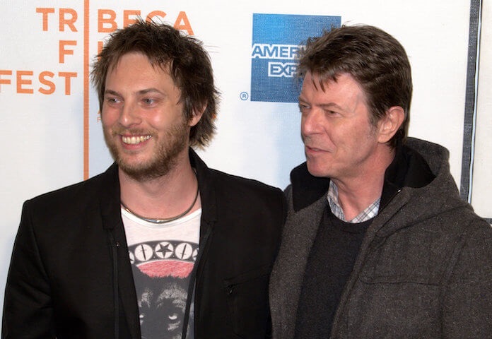 Na foto, da esquerda para a direita, filho de Bowie e ele, sorrindo e olhando para o lado esquerdo. Os dois estão vestidos de preto.
