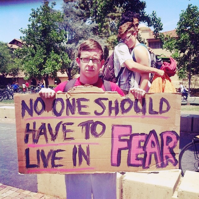 A student holds a sign that says No One Should Have to Live in Fear. Two students embrace in the background.