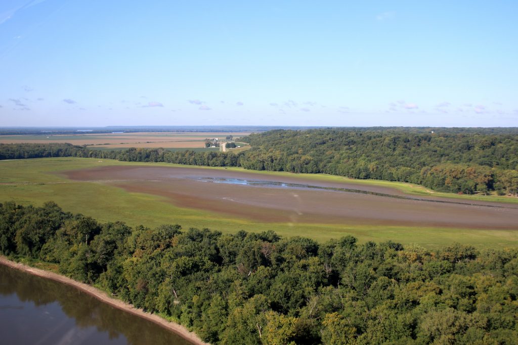 An aerial image of a wetland surrounded by a green field and woodlands. Agricultural fields are in the background with a river in the foreground.