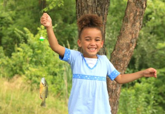 A young girl holds up a fishing line with a small blue-gill fish hooked on line. She is smiling.