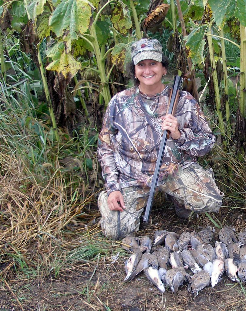 A hunter kneeling on the ground in front of some sunflowers with her harvest of doves on the ground in front of her. She holds her shotgun over her shoulder.