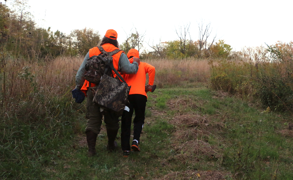 Two hunters in blaze orange hunting gear walking down a path in a prairie. Trees are in the background.