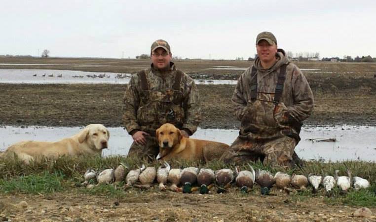 Two hunters in camouflage gear kneeling next to harvested waterfowl with their two hunting dogs. An agricultural field with water standing is in the background.