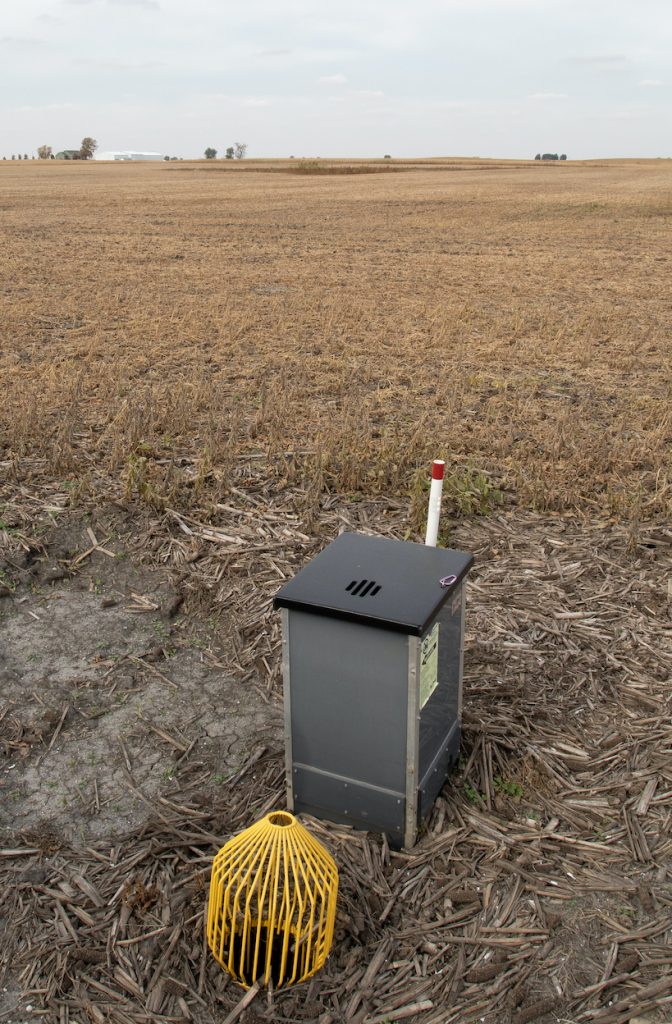 A water control structure in the shape of a metal box with a yellow wire filter over a pipe in a agricultural field during winter.