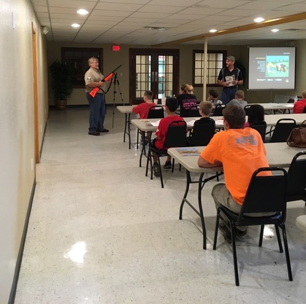 A teacher of hunter safety stands at the front of a classroom and demonstrates the correct way to hold a firearm.