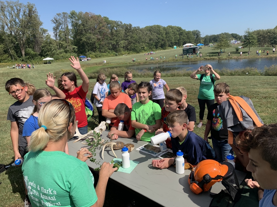 An instructor provides an outreach in the outdoors. A group of children crowd around a rectangular table to participate in the discussion.