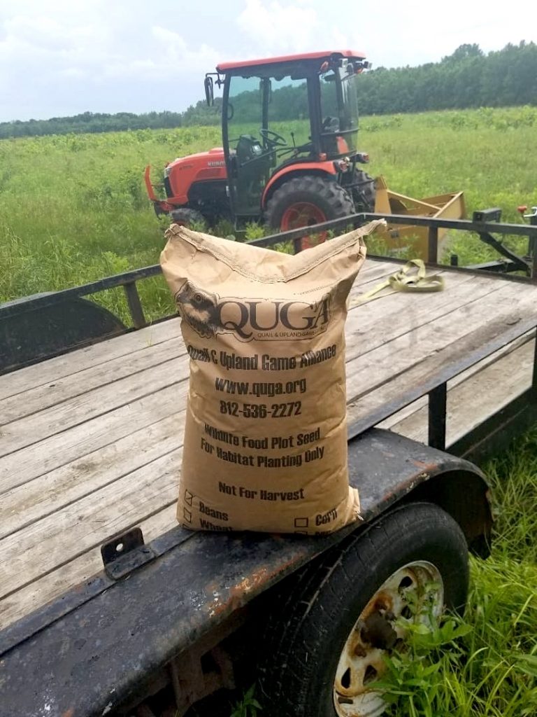 Trailer with a bag of seed sitting on the wheel well is in a grassland. An orange tractor is in the background.