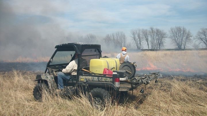 Two men conducting a prescribed burn on a grassland. One man drives an ATV with a water tank. Another man stands outside the vehicle spraying water on the fire line.