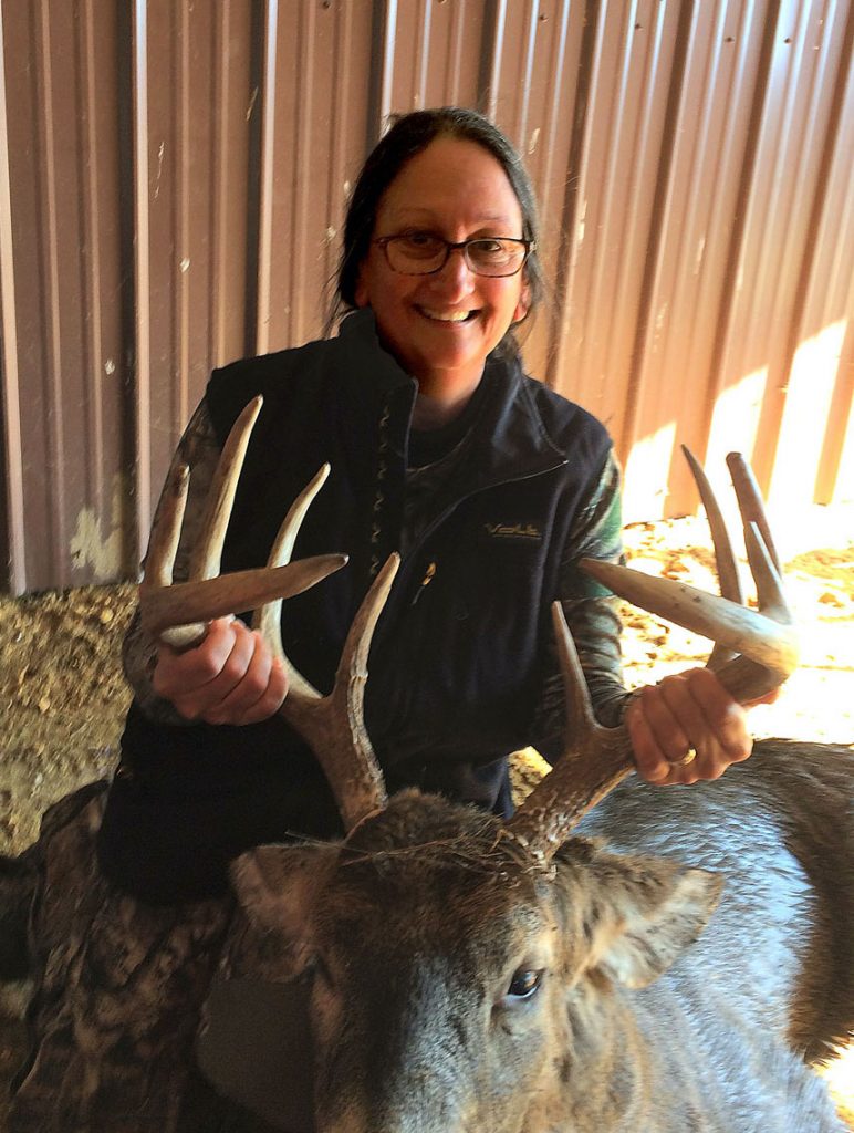 A hunter holds up the antlers of a successful deer harvest. The hunter kneels next to her harvested deer.