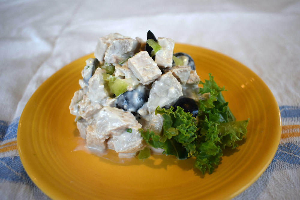 Turkey salad made up of roasted wild turkey breast cubed with diced onion, black olives and mayonnaise with a garnish of green leafy kale is on a bright yellow plate. The plate is resting on a table cloth with a blue and yellow stripe.