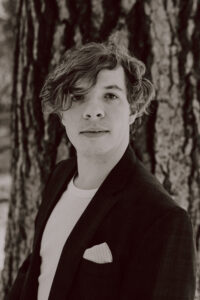[Image description: photo is a black and white headshot of Grant – a white person with long wavy hair. They wear a blazer and the hint of a smile. Background is a gorgeous, blurry Ponderosa pine.]