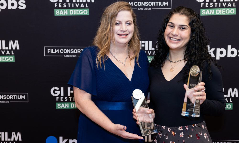 Two women smiling and holding awards in front of a 'GI Film Festival' and 'Film Consortium' backdrop