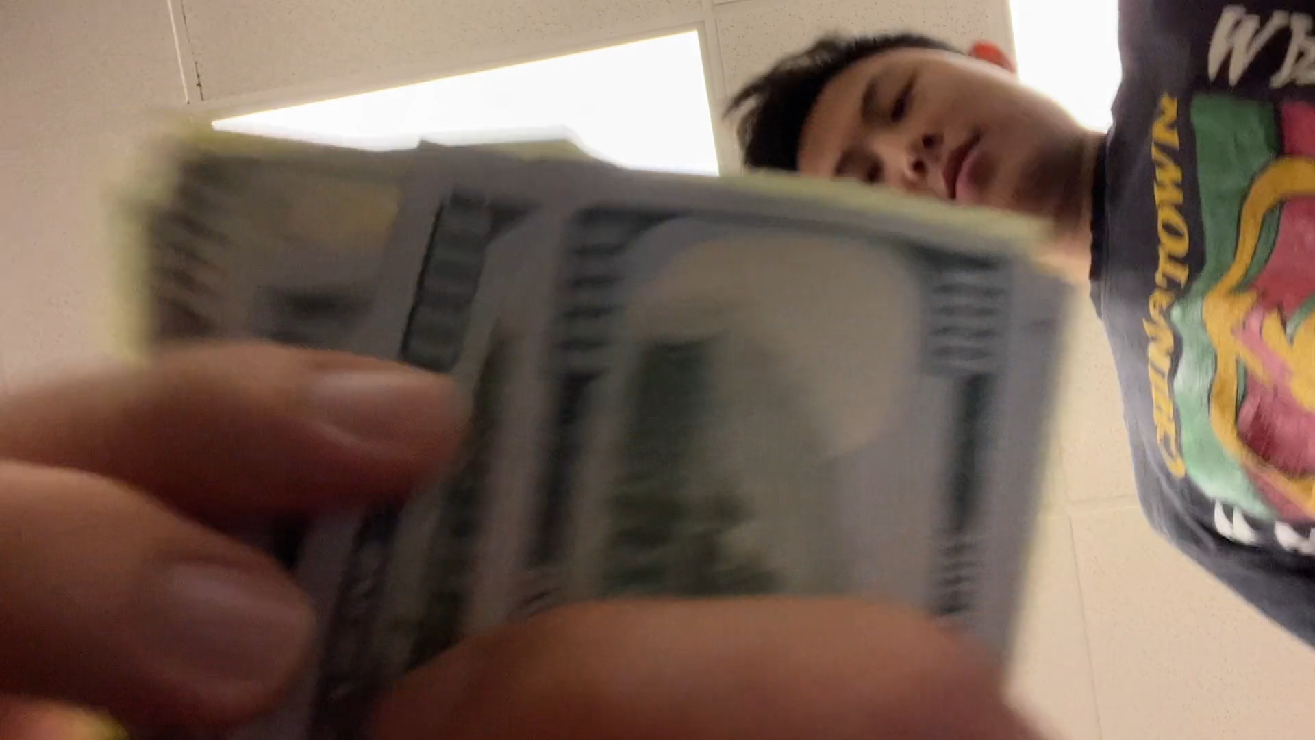 A low-angle shot shows a person looking down while fanning out a stack of 100-dollar bills toward the camera.
