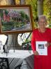 Mary Kocher, holding her book, stands near a painting commissioned by Cindy Gasper, which depicts the Harry Packer Mansion and Gasper's two eldest daughters in the foreground.