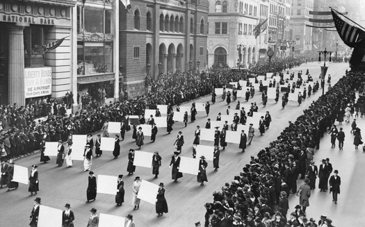 An image of a group of people marching down a street. Several pairs of people are carrying large signs between them. On both sides of the street is a crowd of observers.