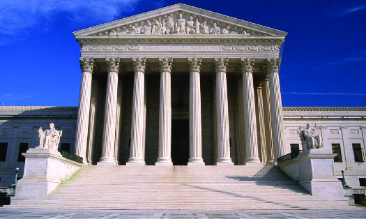 An image of the Supreme Court building. In the foreground, a set of stairs is bracketed by statues on either side, leading up to a portico. The portico has a roof supported by several tall columns.