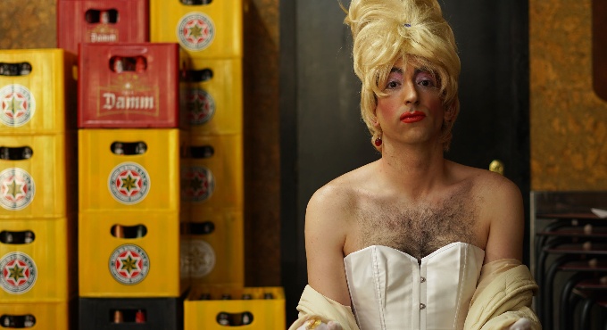 Una persona amb una aparença teatral i andrògina, vestida de blanc, seu sola davant d’un cafè en un bar, destacant amb força enmig d’un entorn quotidià de caixes de cervesa i tons càlids.