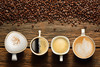four cups of coffee of various shapes and sizes sitting on a table beside a strew of coffee beans
