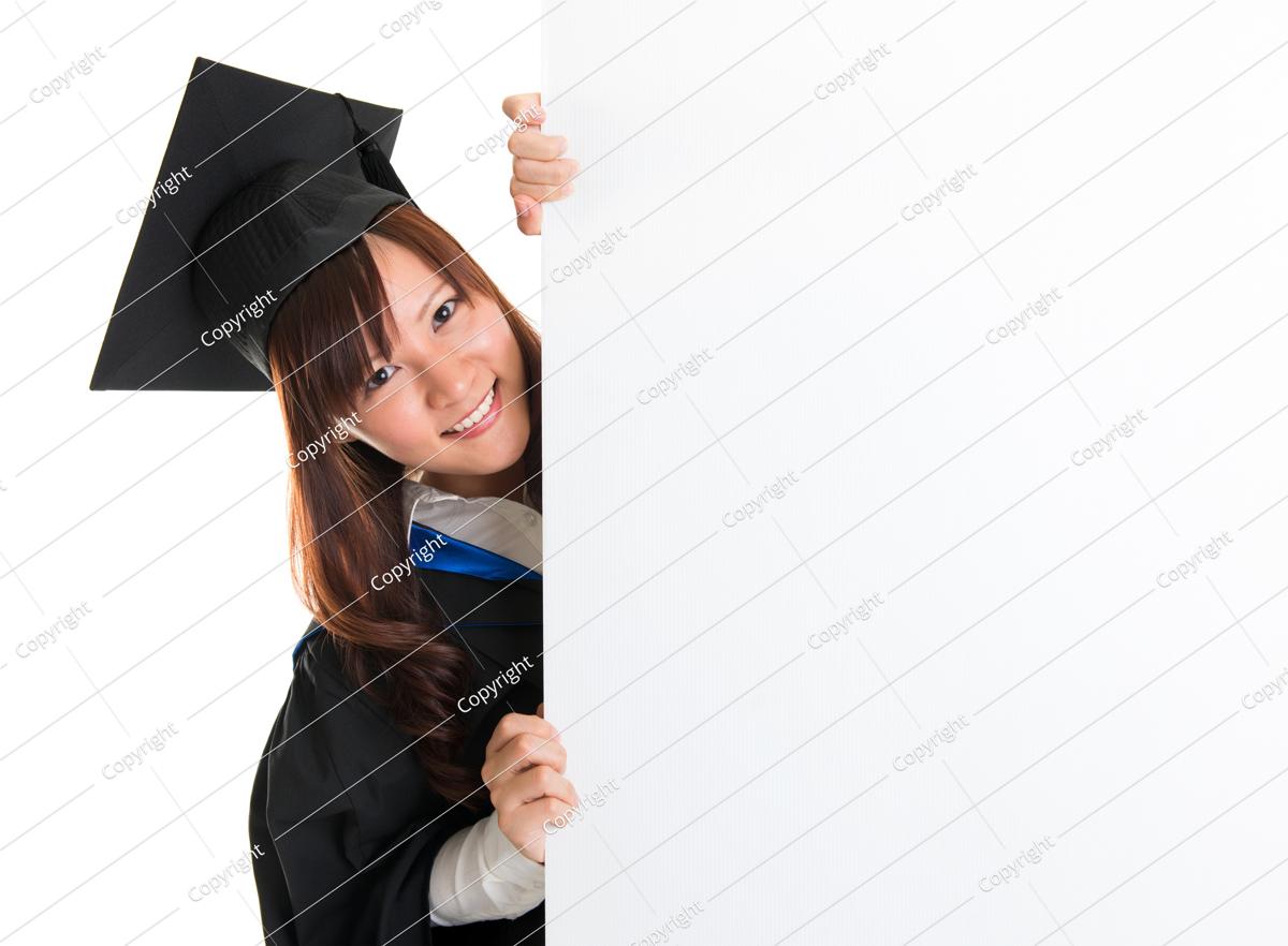 portrait of smiling asian female student in graduate gown peeking, hiding behind a white card, isolated on white background