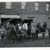 J.E. Keefe wagons with horses & men in front of brick building