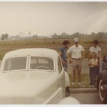 Front of a White Cord with four people between it and another unidentified car, farm in background