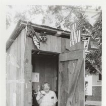 Black and white photo of a Don Aiken sitting in a wooden outhouse