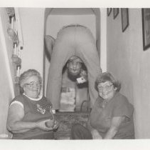 1986 black and white photo of a man bent over, looking back at the camera between his legs while two women laugh while sitting on the stairs in front of him