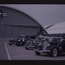1934 Auburn Cabriolet, 1935 or 1936 Auburn Cabriolet, 1928-1930 Auburn, and Cord L-29 next to a plane hangar