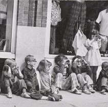 Children sit on the sidewalk along clothing storefronts, one girl standing,