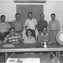 Seven men and one woman standing or sitting in front of a chalk board.