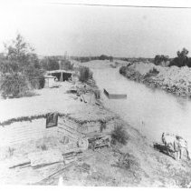 Brush constructed house next to a canal. Man watering horse, flat bottom bo