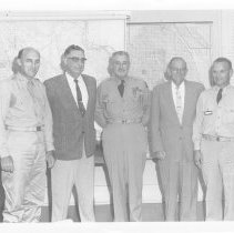 Five men standing in front of wall maps. Harry Free, Herbert Hughes, Elmer