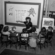 Dayton Heights Elementary School.  Dayton Heights Elementary School activities.  Health program.  Classroom showing background of blackboard exhibits of pictures and charts. [Small group of students listening to teacher’s lesson on what we do to be healthy.]