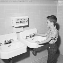Thrift, conservation and school savings pictures. Boy looking at sign on towell [sic: towel] paper rack - "Why use two when one will do."