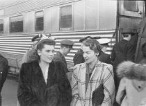 Two women outside passenger train with others in background, saying good-by
