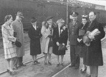 Five women and three men standing outside in front of passenger train, unkn