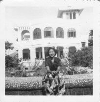 Woman sitting on outside stone fence with house in background, unknown