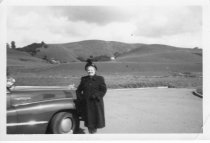 Woman standing next to automobile with hills in background, unknown.