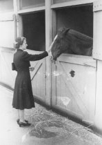 Woman standing next to horse stable touching horse's nose, unknown.