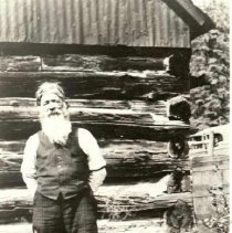 Potato Creek Johnny in front of log cabin.
