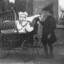 K-81-13-10 Child posing next to a baby in a wicker carriage.