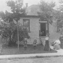 K-81-13-18 People posing in front of a small brick house, by the sidewalk.