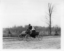 Black and white street view photograph of a father and child automobiling in a horseless carriage along a dirt road