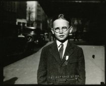 Black and white enlargement of a portrait of a boy wearing glasses, a tie, a jacket, and a safety patrol medal standing outside the entrance to the Statler Hotel on Bagely Street
