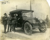 Black and white photographic print depicting three men standing next to a parked automobile with a boy sitting behind the wheel