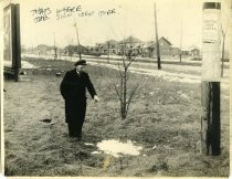 Black and white photographic print depicting a man pointing to stump in the ground where a signpost had been