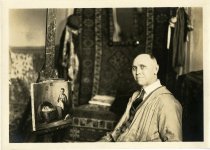 Sepia-toned portrait photo of Charles Waltensperger posed seated in his studio in front of a painting of a woman sewing beside a baby